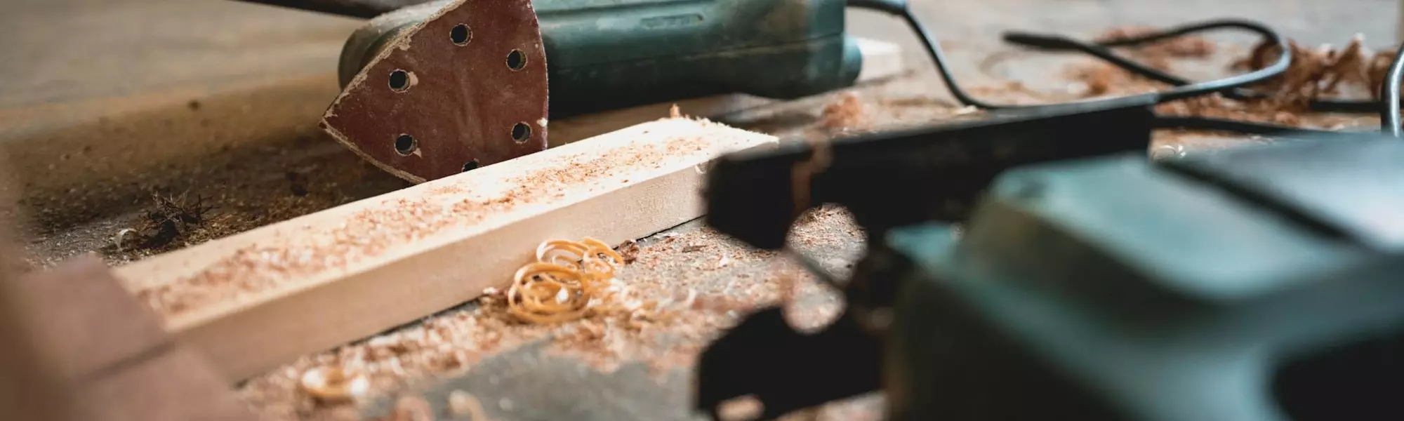 Detailaufnahme von Schleifgerät und Stichsäge auf Arbeitstisch mit Holzlatte und Holzspänen